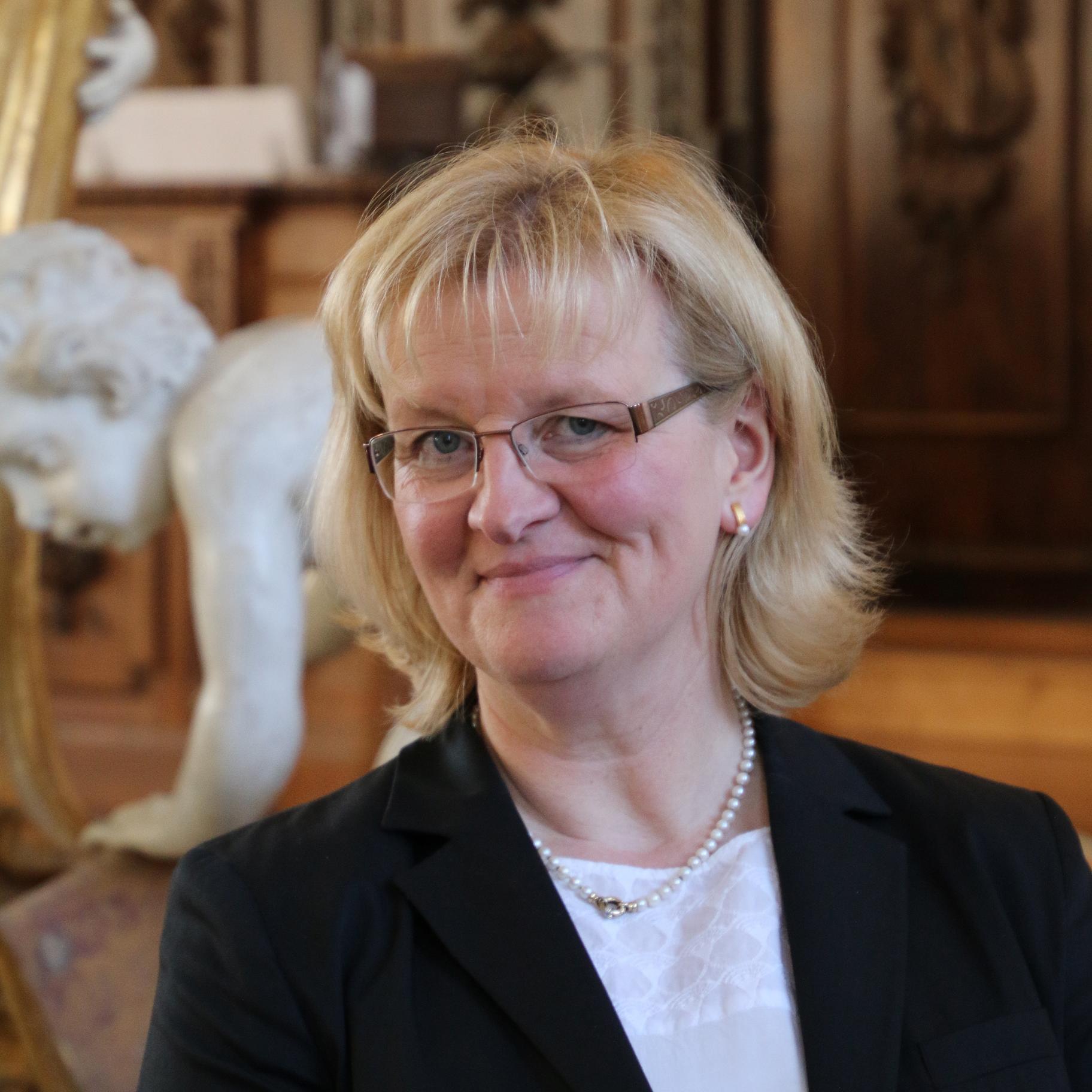 Frau mit blonden, halblangen Haar, Brille, weißer Bluse, schwarzer Jacke