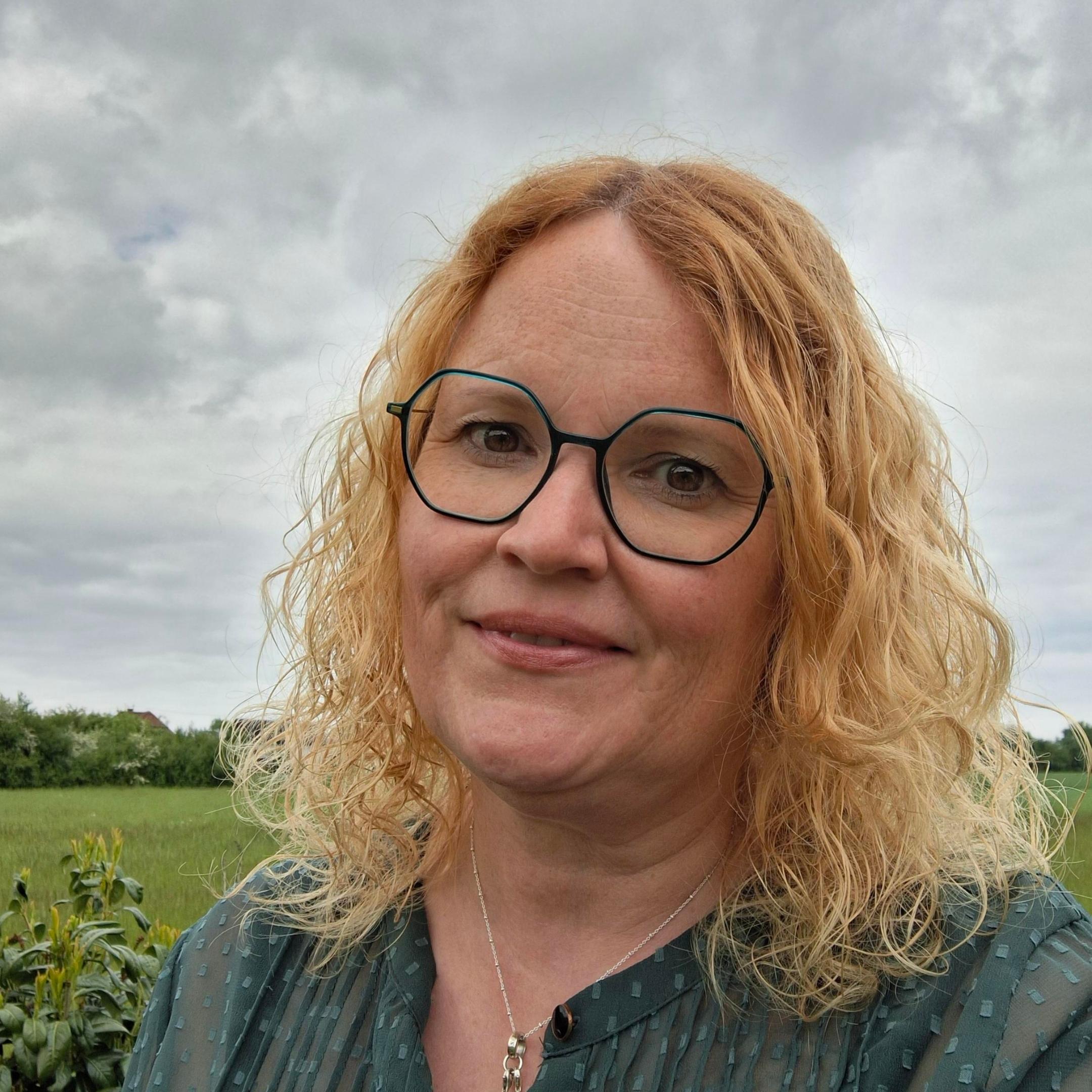 Frau mit blonden, halblangen Locken, grüner, schmaler Brille
